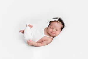 Sleeping newborn baby girl in white knit wrap with bow headband on white background.