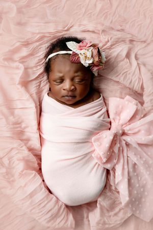 Professional newborn photographer in Menifee: Baby girl swaddled in pink wrap with floral headband sleeping on pink blanket.