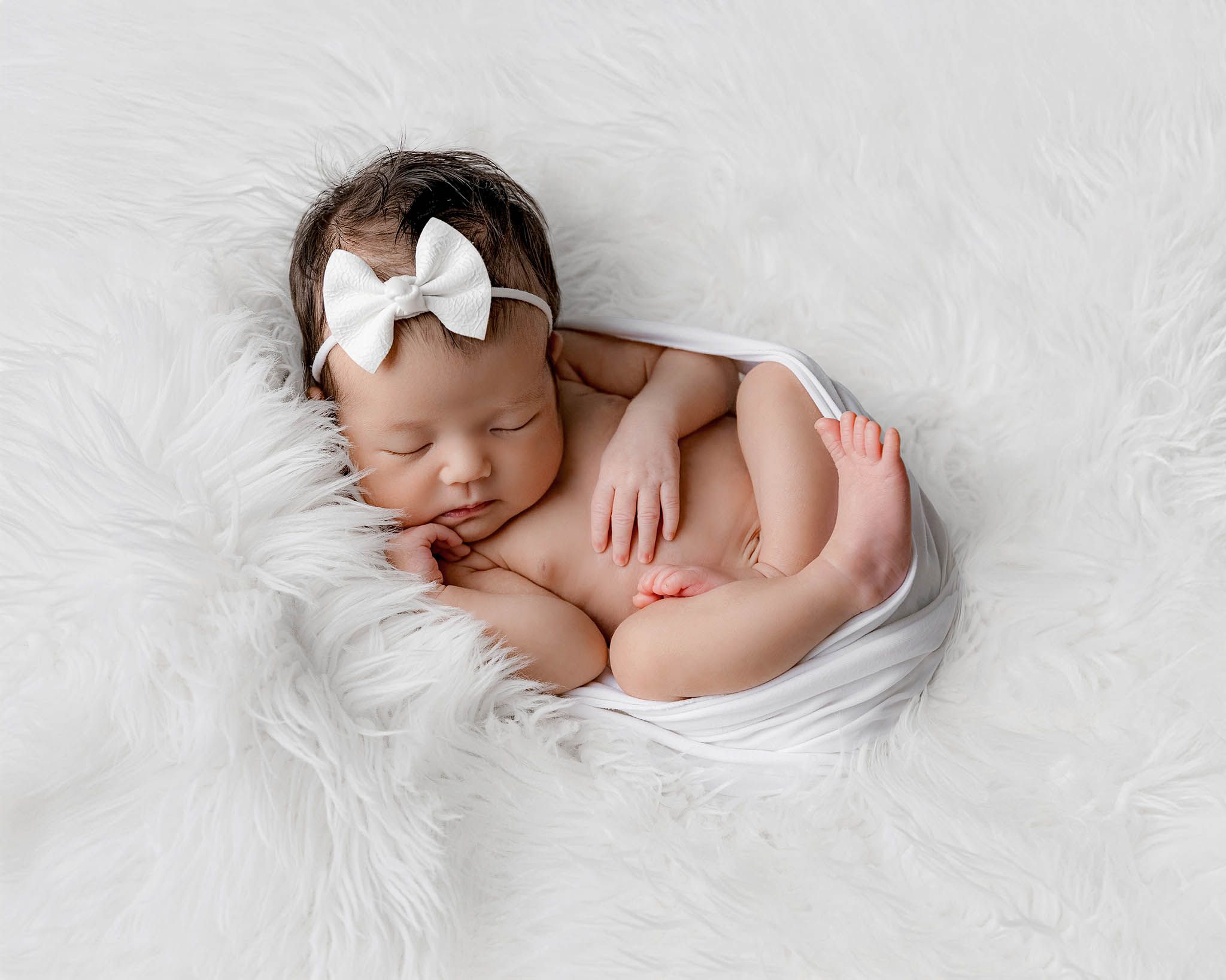 Sleeping newborn baby girl with white bow headband posed on fluffy white fur blanket.