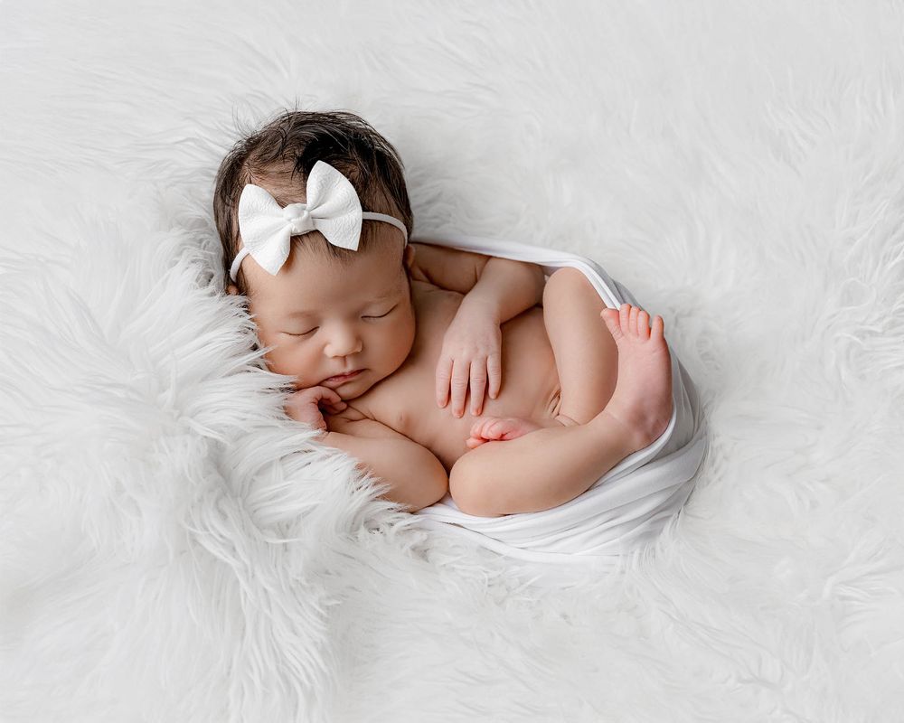 Sleeping newborn baby girl with white bow headband posed on fluffy white fur blanket.