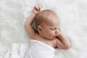 Sleeping newborn baby wrapped in white swaddle lying on soft white fluffy fur blanket.
