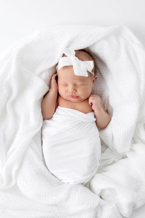 Newborn baby girl swaddled in white wrap with white bow headband sleeping on white blanket.