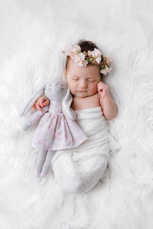 Sleeping newborn baby girl with floral crown holding stuffed bunny toy on white fur blanket.