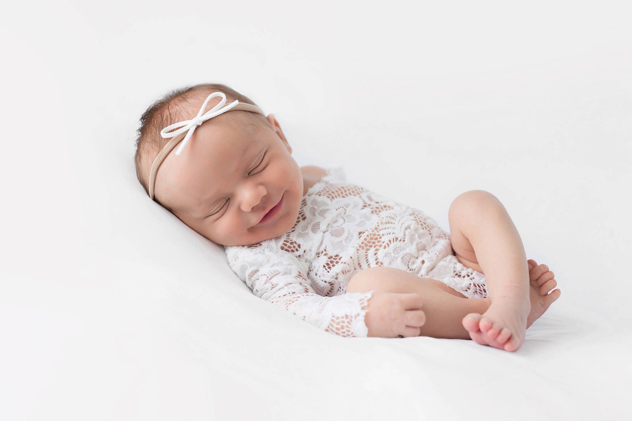 Smiling newborn baby girl in white lace romper and bow headband lying on white background.