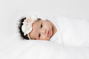 Newborn baby girl swaddled in white knit wrap wearing floral headband looking at camera.
