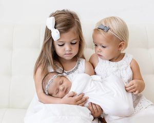 Two sisters in white dresses holding their sleeping swaddled newborn baby sister.