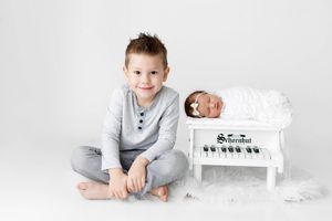 Newborn sibling photographer: Smiling little boy sitting beside sleeping baby sister in white studio.