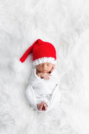 Swaddled newborn baby sleeping on white fur wearing red Santa hat.