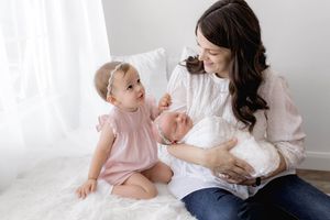 Newborn family photographer: Mom smiling at toddler daughter while holding sleeping newborn baby in lifestyle photoshoot.