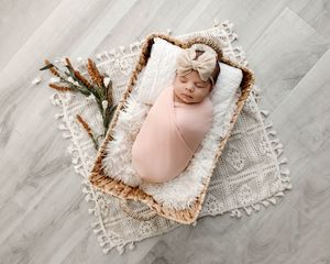 Swaddled newborn in peach wrap sleeping in wicker basket on boho rug.