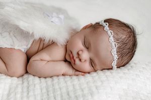 Sleeping newborn baby girl with lace headband and white angel wings on blanket.