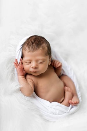 Sleeping newborn baby wrapped in soft white blanket on fluffy white surface.