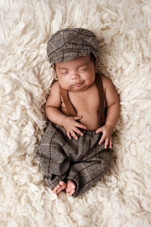 Adorable newborn baby sleeping in vintage tweed outfit with suspenders and newsboy cap on fluffy rug.