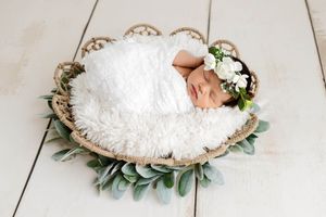 Sleeping newborn baby wrapped in white swaddle with floral crown resting in a wicker heart basket.
