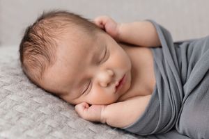 Sleeping newborn baby boy swaddled in gray wrap resting on soft textured blanket.