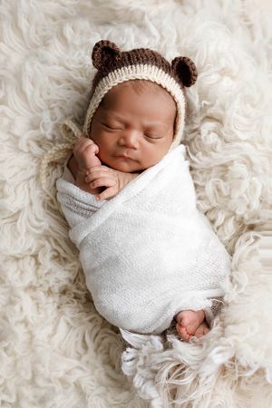 Newborn baby boy swaddled in white wrap wearing a knit bear ear hat on a cream fur blanket.