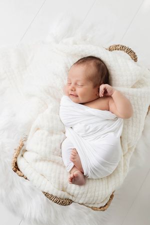 Smiling newborn baby swaddled in white wrap sleeping peacefully in a wicker Moses basket.