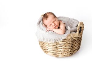 Sleeping newborn baby nestled in gray fleece inside a round wicker basket on white background.