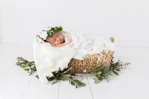 Sleeping newborn baby in white lace wrap with flower crown in wicker basket with greenery.