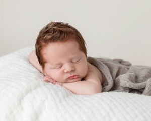 Sleeping newborn baby with red hair resting chin on hands on white textured blanket.