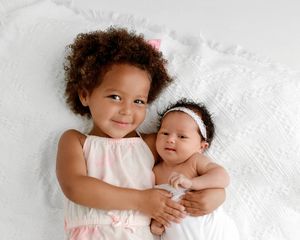 Smiling toddler girl gently holding her newborn baby sibling on a soft white blanket.
