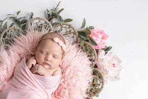 Sleeping newborn baby girl wrapped in pink swaddle with floral headband in a wicker basket with pink flowers.