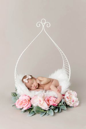Newborn baby girl sleeping in a decorative white metal swing surrounded by pink peonies and roses.