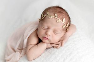 Sleeping newborn baby wearing a delicate gold leaf headband wrapped in a soft pink blanket.