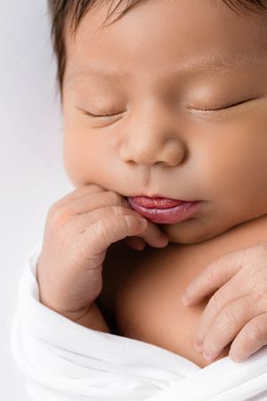 Close-up portrait of a sleeping newborn baby wrapped in white with tiny hands near face.