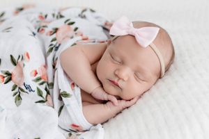Newborn baby girl sleeping peacefully wrapped in a floral muslin swaddle with a pink bow headband.