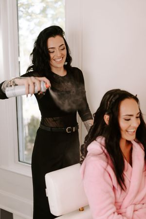 Hair stylist in black dress spraying product on smiling woman in pink robe during bridal preparation.