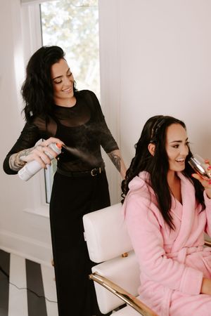 Hair stylist in black outfit spraying hairspray on brunette bride in pink robe during wedding preparation.