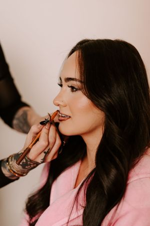 Tattooed makeup artist applying lip liner to a young woman with long dark wavy hair.