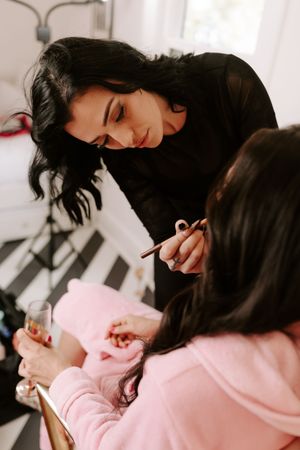 A makeup artist in black applies makeup to a woman in a pink hoodie holding a champagne glass.