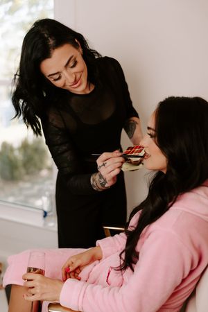 makeup artist applying lipstick to brunette bride in pink robe holding champagne glass.
