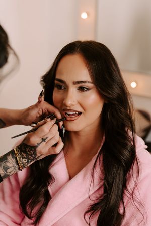 Close-up of brunette bride in pink robe getting lipstick applied by tattooed makeup artist.