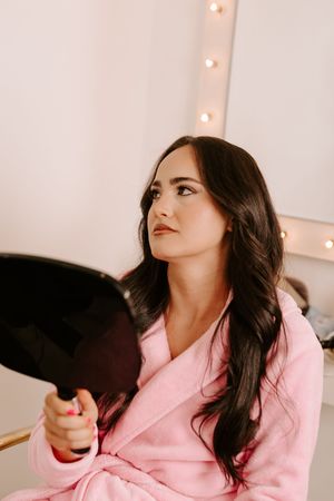 Woman in pink robe holding mirror, gazing upward near vanity with warm lights.