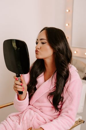 Woman in pink robe blowing kiss at handheld mirror in bright vanity room with Hollywood lights.