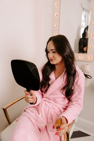 A woman in a pink fluffy robe sits examining her reflection in a handheld mirror in a vanity room.