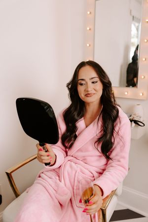Woman in pink robe holding hand mirror in vanity room with Hollywood lights, getting ready with pink manicure.