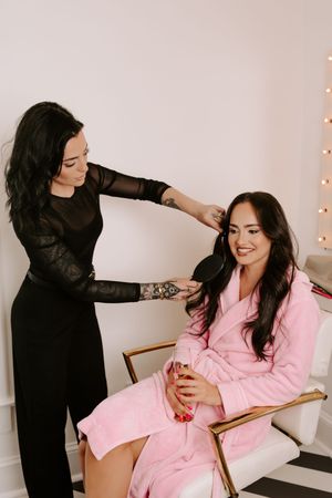 Hair stylist in black dress styling brunette woman in pink robe holding champagne in a bright salon.