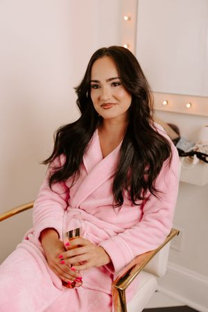 Woman in pink robe holding champagne glass, seated in gold chair near vanity mirror with lights.