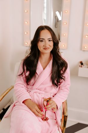 Woman in pink robe sitting in glam vanity room holding champagne glass with warm bulb mirror lights behind her.