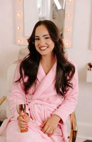 Smiling woman in pink robe holding champagne flute, seated in front of lit vanity mirror in bright salon.