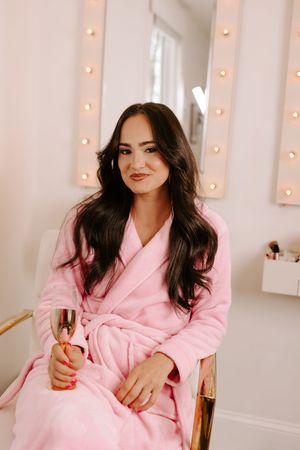 Woman in pink robe holding rose gold champagne glass seated in glamorous vanity room with lit mirror.