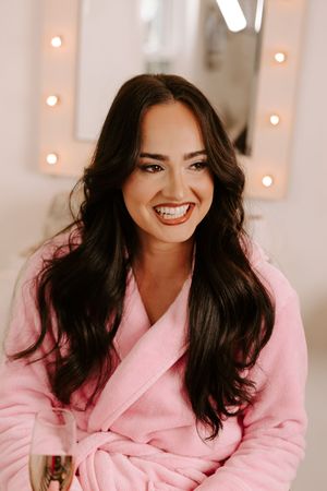 Smiling woman with long dark hair wearing a pink fluffy robe, sitting in front of a lit vanity mirror.