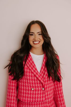 Smiling woman with long dark hair wearing a pink houndstooth blazer over a white top against a light background.