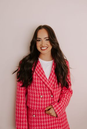 Smiling woman with long dark hair wearing a pink houndstooth blazer over a white top against a light background.