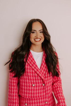 Smiling woman with long dark hair wearing a pink houndstooth double-breasted blazer over a white top.