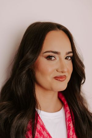 Young woman with long dark hair, red lips, and pink scarf poses against a white background.